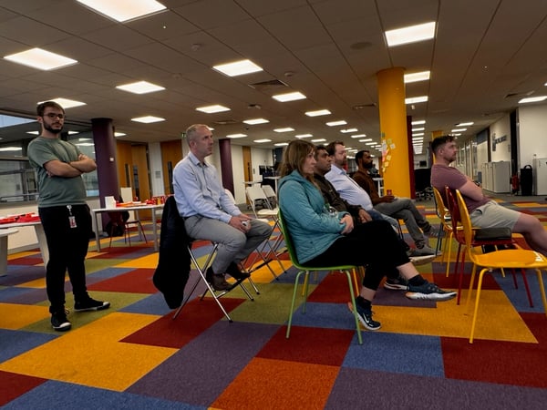 A group of six people is gathered indoors in a modern office space, sitting and standing while attending a presentation or meeting, characterized by a brightly colored, geometric carpet.