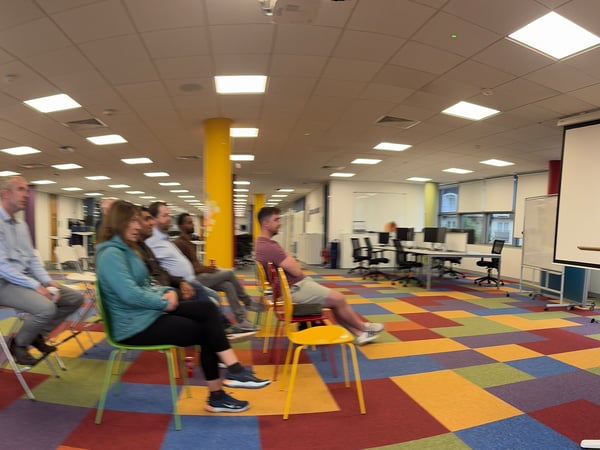 A group of people is attending an indoor presentation or meeting in a modern, brightly lit office space characterized by a striking, multi-colored checkered carpet.