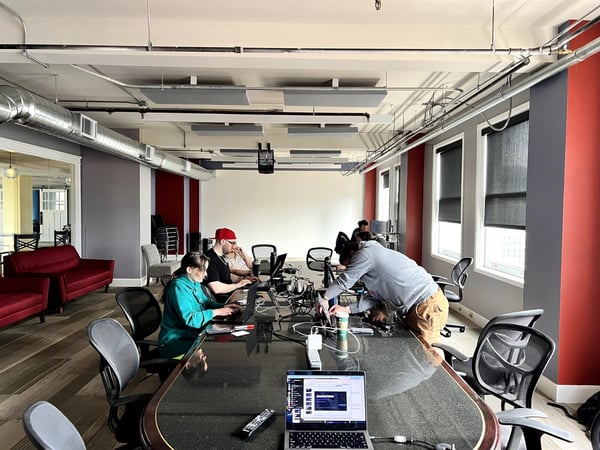 A group of people are working on laptops around a large, dark conference table in a modern office space with industrial-style exposed ceilings.