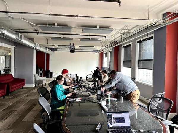A group of people are working on laptops around a large conference table in a modern office space with industrial ceilings.