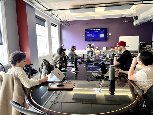 A group of people are seated around a large black conference table in a modern office, attending a presentation shown on a wall-mounted screen.