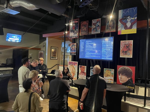 A group of people is gathered inside a dimly lit venue, decorated with music posters, observing a large monitor displaying computer code or terminal output, likely during a technical demonstration or event.