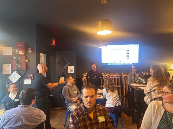 A group of people are gathered in a dimly lit room with dark walls, attending a presentation or social event where a man is speaking in front of a screen and wine racks.