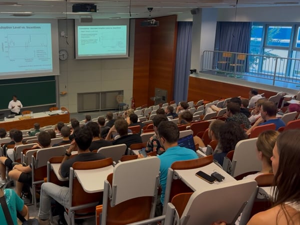 A photograph capturing a lecture or presentation taking place in a large, tiered auditorium filled with a seated audience facing a speaker and a projected slide.