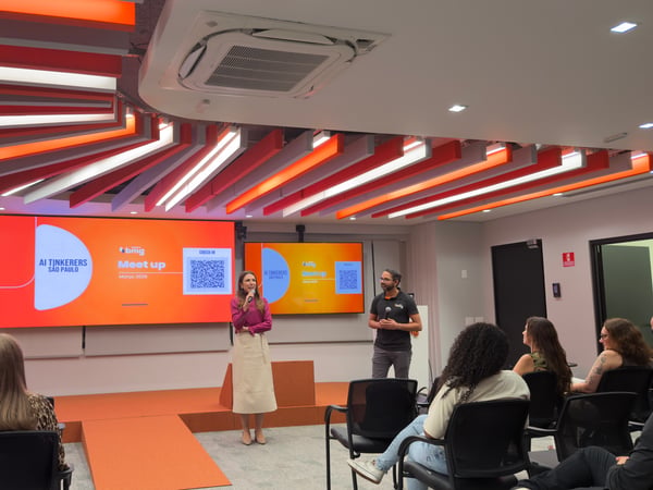 A woman speaks into a microphone on a stage during an 'AI Tinkerers São Paulo' meetup event, with an audience seated in the foreground.