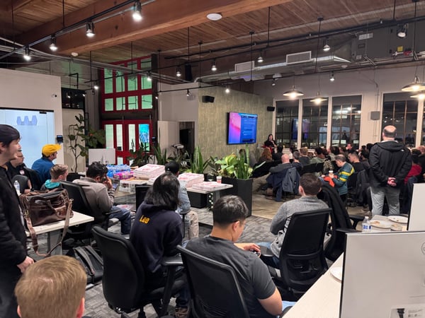 A photograph captures a large group of people attending an indoor presentation or meetup in a modern office space featuring exposed wooden beams and industrial lighting. A presenter stands near a large screen displaying slides, while the audience is seated, mostly facing away from the camera.