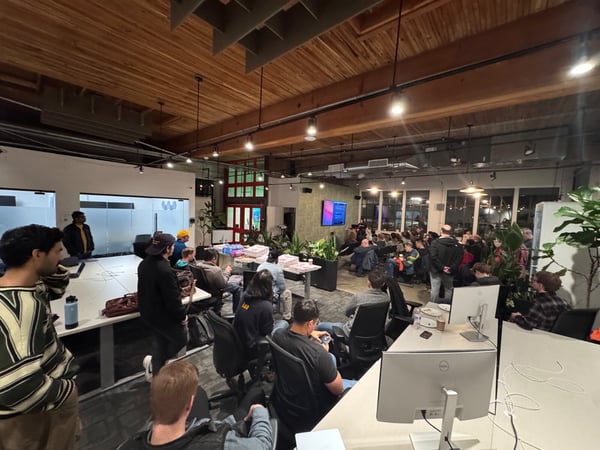 A candid photo captures a large gathering of people attending a presentation or meeting inside a modern, loft-style office featuring exposed wooden ceilings and large windows. Attendees are seated and standing, mostly facing a screen visible in the distance.