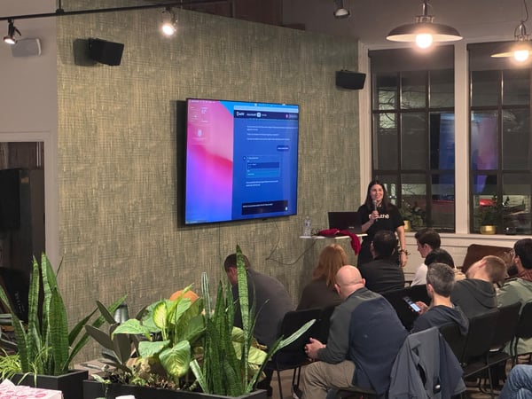 A photograph capturing an indoor presentation or tech talk, showing a woman speaking into a microphone in front of a large screen displaying a user interface, while an audience is seated and listening.