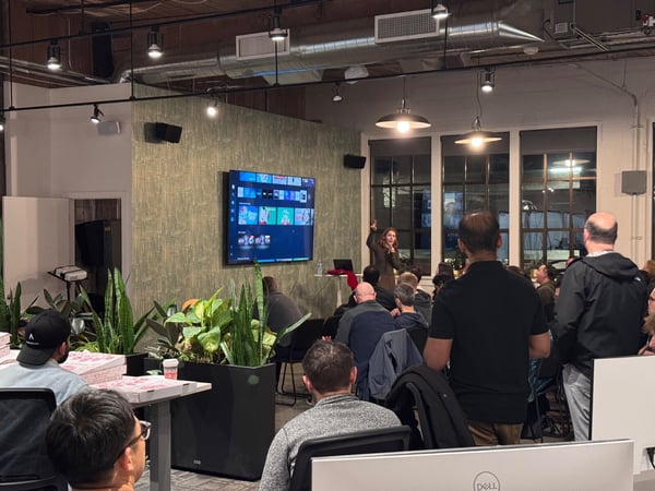 A photograph capturing an indoor event where a woman is giving a presentation to a large, seated audience in a modern office space with industrial design elements and exposed ductwork.