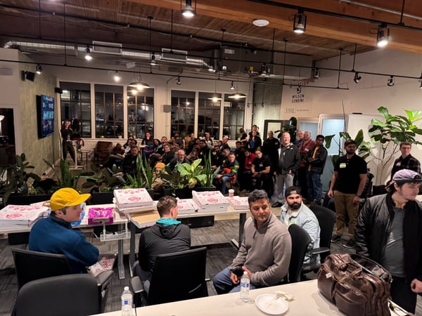 A wide-angle photograph captures a large crowd of people attending an evening event or meetup in a modern, industrial-style indoor space, with several pizza boxes visible on tables in the foreground.