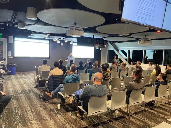 A wide-angle photo capturing an indoor presentation or lecture, showing an audience seated in rows facing a presenter at a podium and two large projection screens.
