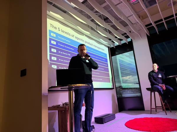 A man is giving a presentation on a stage about 'The 5 levels of agentic product management' while another man sits on a stool nearby.