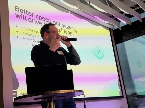 A man stands on a stage giving a presentation with a microphone in hand, positioned in front of a large screen displaying a slide about coding agents.