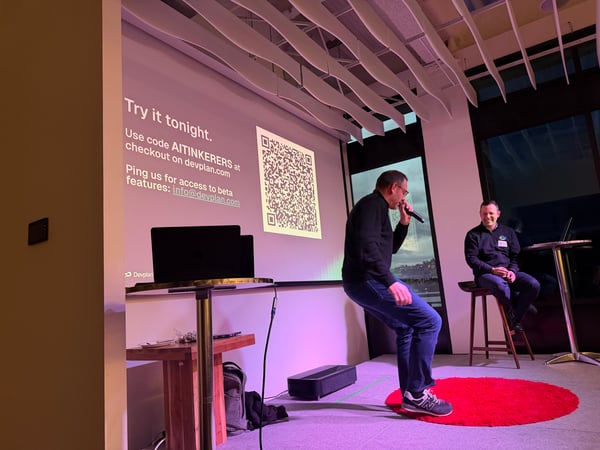 A man stands on a stage speaking into a microphone during a presentation, with a projection screen behind him displaying a QR code and promotional text. Another man sits on a stool to the side, observing the speaker.