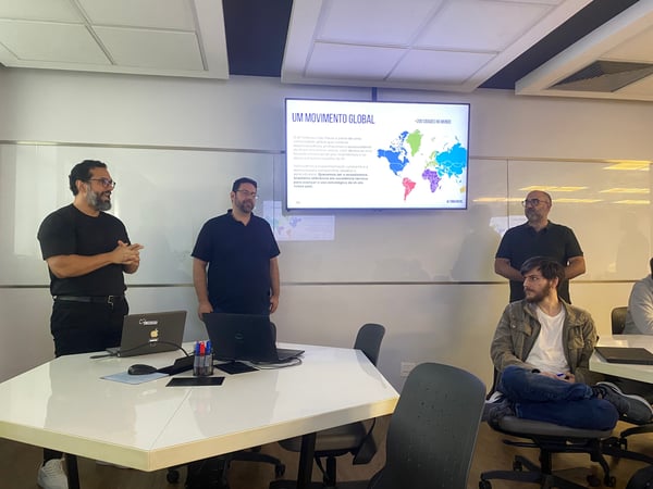 A group of men in an office setting attending a presentation about 'AI Tinkerer São Paulo', featuring a screen with a world map and Portuguese text.