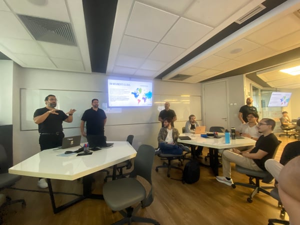 A man is giving a presentation to a group of people in a modern office meeting room, with a screen displaying a map and text in the background.