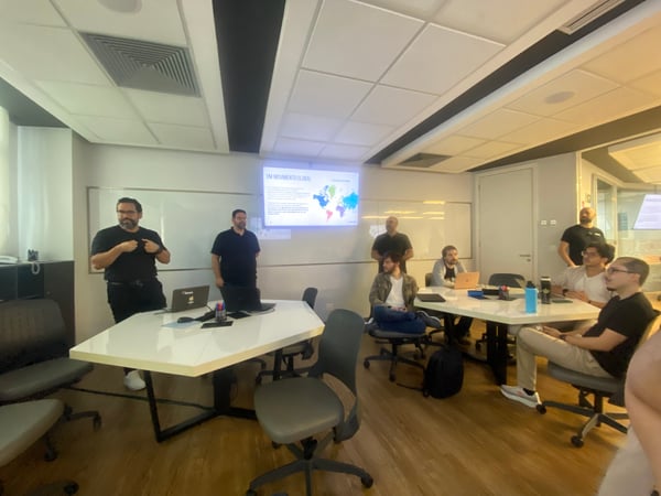 A group of men are gathered in a modern office meeting room for a presentation, with one man standing and gesturing towards a screen displaying a slide titled 'UM MOVIMENTO GLOBAL'.