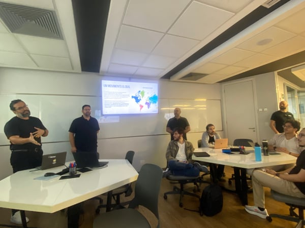 A group of men in an office setting attending a presentation. One man is standing and gesturing while a screen displays a slide titled 'UM MOVIMENTO GLOBAL' with a colorful world map.