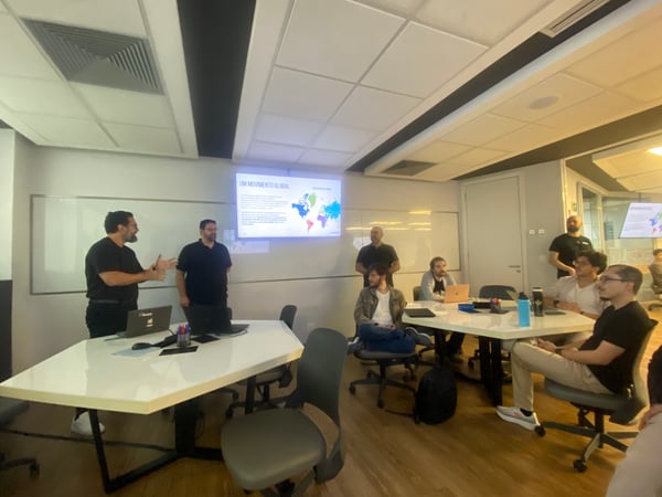 A group of men in a modern office setting attending a presentation titled 'UM MOVIMENTO GLOBAL' shown on a wall-mounted screen.