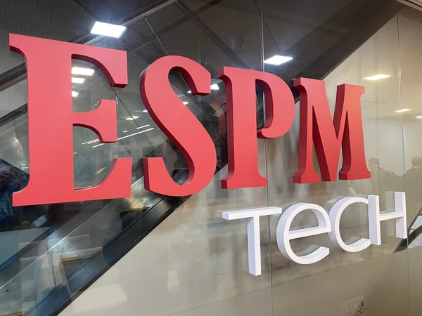 A photograph showing a large 3D logo for 'ESPM tech' mounted on a glass wall inside a modern building.