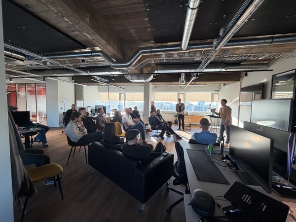 A group of people gathered in a modern, industrial-style office for a presentation or meeting, with one person standing and speaking to the group.