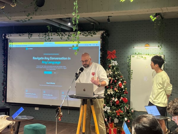 A photograph capturing an indoor presentation where a man is speaking into a microphone while displaying a website titled 'Local Guide' on a large projection screen. The room is decorated with Christmas items, including a large tree, and artificial ivy hangs from the ceiling.