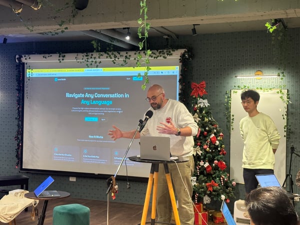 A photograph capturing an indoor presentation or tech demo, where a man in a light sweater speaks into a microphone, gesturing toward a large projection screen displaying a website titled 'Navigate Any Conversation in Any Language.' A second man stands to the right, adjacent to a heavily decorated Christmas tree.