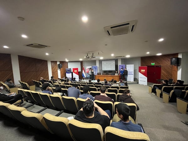A wide-angle photograph of a presentation or seminar taking place in an indoor auditorium with tiered seating and a stage featuring several presenters and banners.