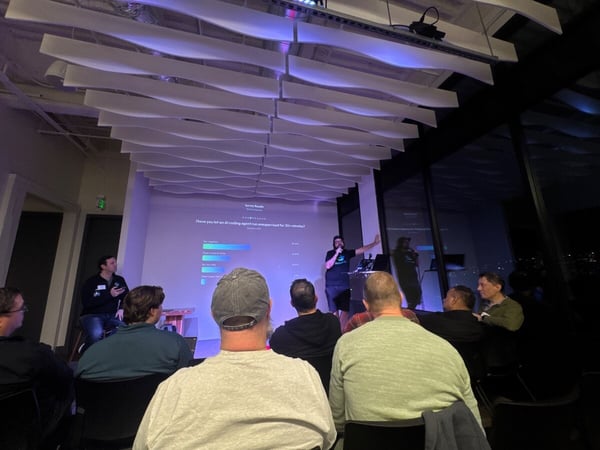 A nighttime photograph captures an indoor presentation event where an audience is seated, watching two speakers in front of a projection screen displaying survey results about AI coding agents. The venue features large windows overlooking a city skyline and a distinctive wavy ceiling structure illuminated with purple light.