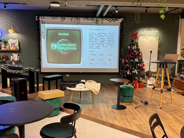 A photo of a modern, well-decorated indoor event space, likely an office or co-working area, set up for a hackathon and featuring Christmas decorations, with event details projected onto a large screen.