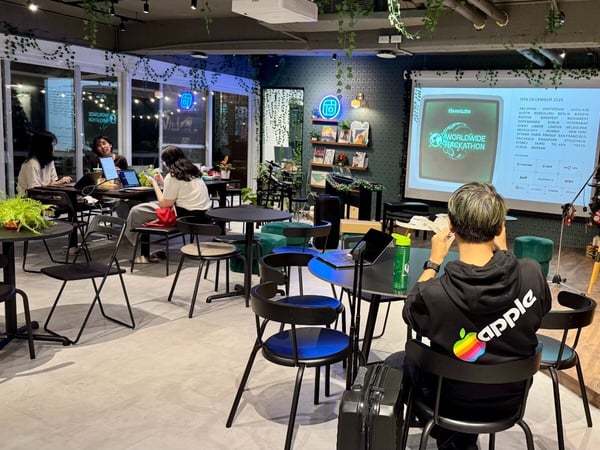 A candid photograph captures an indoor event or co-working session where several individuals are seated at tables using laptops, with a large screen displaying details for a "WORLDWIDE HACKATHON" event in a room decorated with plants and string lights.