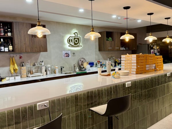 A photograph showing the interior of a modern communal kitchen or bar area, featuring a long white countertop and a facade clad in dark green vertical tiles.