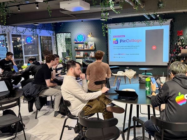 A photograph capturing an indoor presentation or workshop where a group of people are seated around tables, mostly looking toward a projection screen displaying information about the 'PicCollage' product, while a woman stands presenting on the right.