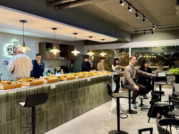 A photograph capturing a casual social gathering or office event where a group of people are serving themselves pizza from numerous boxes laid out on a long, tiled bar counter in a modern, well-lit interior space. Several attendees are seated at nearby high tables, some appearing to be working on laptops or engaged in discussion.