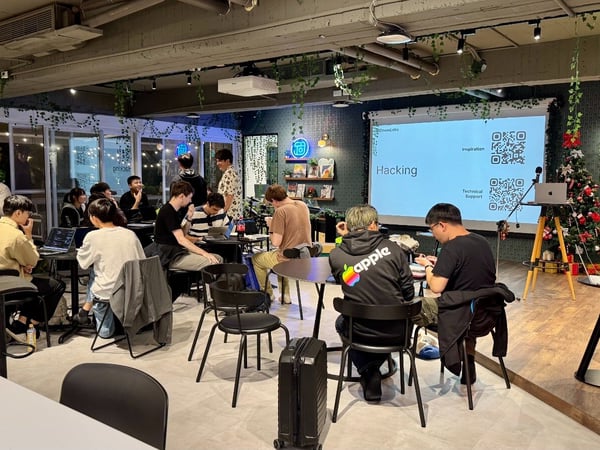 A candid photograph capturing an indoor hackathon or workshop event where a large group of people are seated at tables, intensely working on open laptops. A projector screen in the background displays the title "Hacking" along with QR codes for inspiration and technical support.