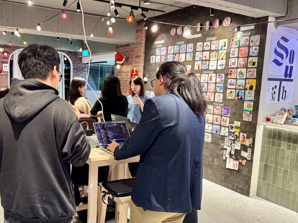 A candid photograph captures a group of six people gathered around a high table in a warmly lit indoor workspace decorated with Christmas accents and featuring a large wall display of small square photos/cards.