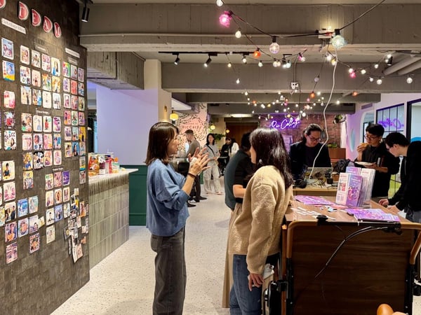 A candid photograph capturing a casual indoor gathering or event, featuring several people interacting near a table and a large wall covered in small photo collages. String lights hang overhead, illuminating the modern, warmly lit space.