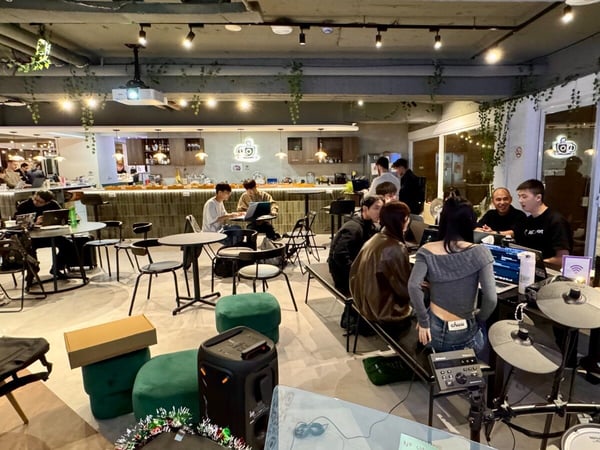 A candid photograph capturing a group of people working and socializing in a modern, dimly lit indoor co-working space or cafe, featuring laptops, small tables, and an electronic drum kit in the foreground.