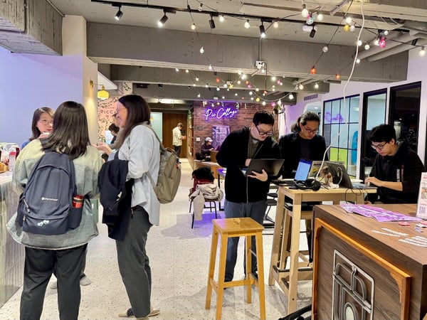 A candid indoor photograph capturing a group of people at what appears to be a casual event or co-working session, with some socializing near a counter and others working intently on laptops at high tables.