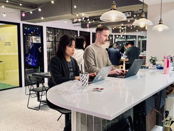Two people are seated at a long white counter, focused on working on their open laptops in a modern, industrial-style co-working space featuring string lights and a 'GAME ROOM' sign on a yellow wall.