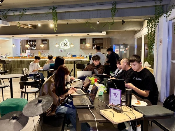 A candid photo showing a group of people working intensely on laptops at tables within a modern, cafe-like co-working space decorated with hanging plants and featuring a bar area in the background.