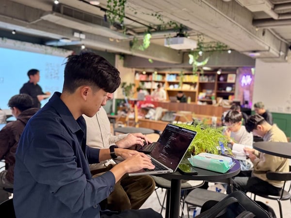 A candid photograph capturing a group of people working intensely on laptops during what appears to be a hackathon or coding event, set in a modern indoor venue decorated with plants and exposed infrastructure.