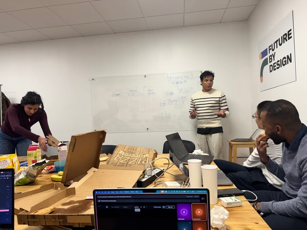 A group of people are in a meeting room with one person standing and gesturing towards a whiteboard filled with diagrams, while others sit at a table with laptops and pizza boxes.