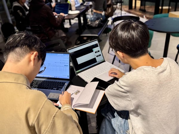 A candid photograph shows two people sitting closely at a table, working intently on their open laptops and notebooks in a dimly lit indoor setting, likely a shared workspace or cafe.