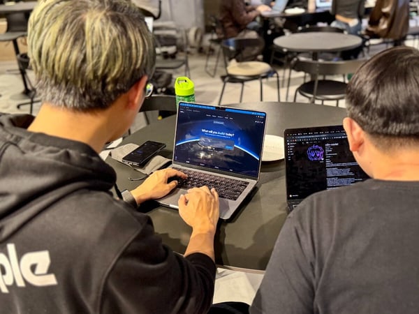 A candid photo shows two individuals working on separate laptops at a dark table in an indoor communal workspace, focusing intently on their screens, suggesting a coding or development session.