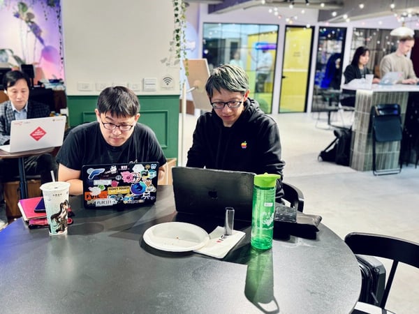 A candid indoor photograph capturing five people working on laptops in a modern co-working space or office lounge featuring tiered wooden seating and colorful glass partitions.