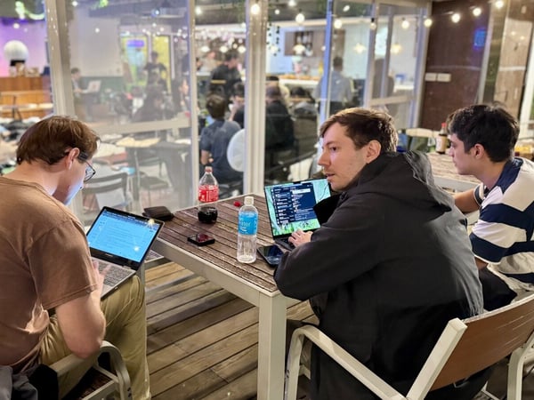Three men are sitting at an outdoor or semi-outdoor table working on open laptops during the evening, with a busy, brightly lit indoor space visible through glass windows behind them.