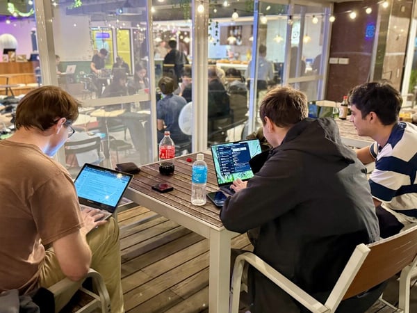 Three young men are seated at a slatted outdoor table, focused on their open laptops during an evening work session, with an illuminated indoor communal space visible through glass doors behind them.