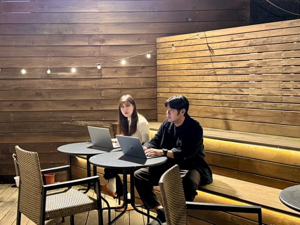 A candid photo capturing a man and a woman sitting at small tables on a wooden outdoor patio at night, both focused on working on their open laptops under the soft glow of string lights.