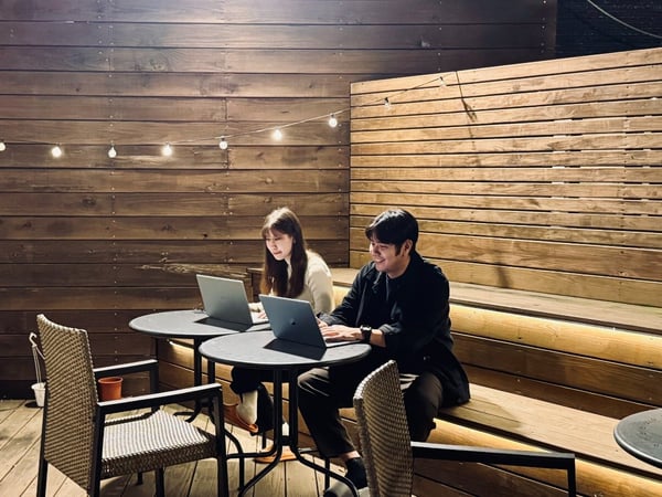 Two people, a man and a woman, are sitting at small round tables working on open laptops on a dimly lit outdoor wooden deck or patio at night.